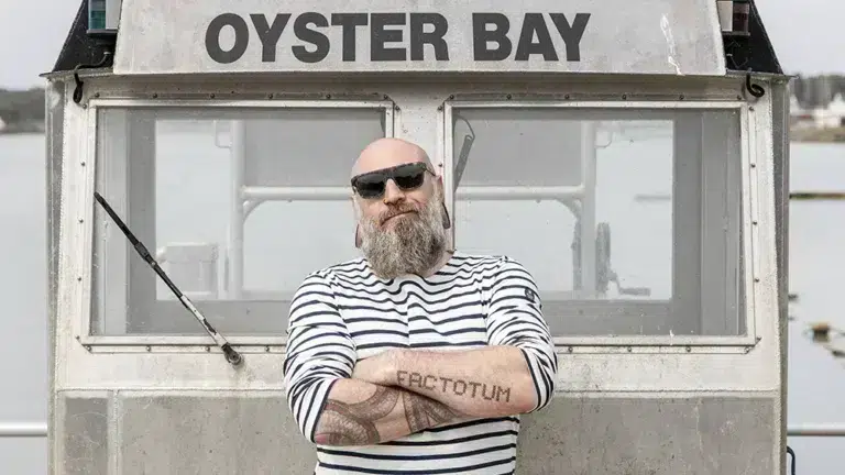 Homme barbu portant des lunettes de soleil Friendly Frenchy devant un bateau Oyster Bay, illustrant l'engagement durable et solidaire de la marque.
