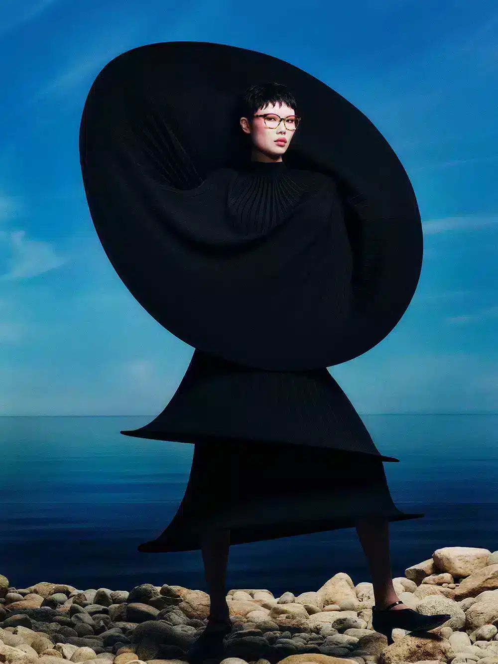 Mujer con gafas ópticas de la colección SS25 de Etnia Barcelona, con montura roja y estilo vanguardista, frente a un paisaje marino.