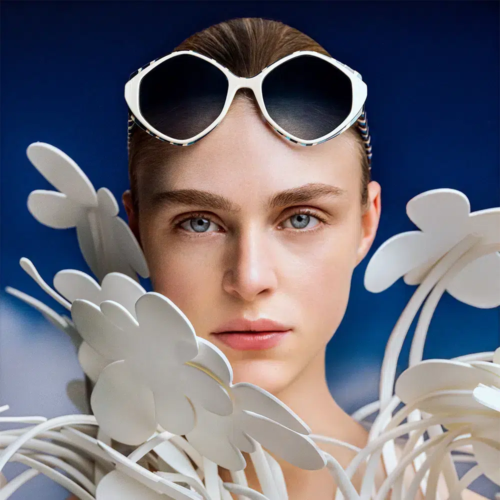 Portrait of a woman wearing white sunglasses from Etnia Barcelona’s SS25 collection, surrounded by embossed floral patterns.