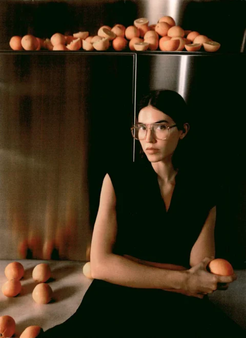 Femme portant des lunettes de vue KALEOS dans un cadre minimaliste avec des pêches, tiré du lookbook Printemps-Été 2025.