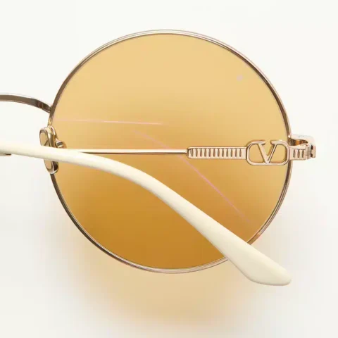 Valentino Eyewear, a close-up of the glasses featuring a gold metal frame, VLogo Signature, and amber-tinted lenses, from the Avant les Débuts collection by Alessandro Michele.