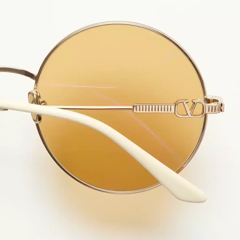 Valentino Eyewear, a close-up of the glasses featuring a gold metal frame, VLogo Signature, and amber-tinted lenses, from the Avant les Débuts collection by Alessandro Michele.