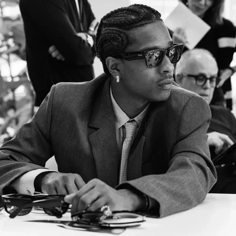A$AP Rocky x Ray-Ban. A$AP Rocky in a gray suit and Ray-Ban glasses, carefully observing a selection of frames during a creative meeting.