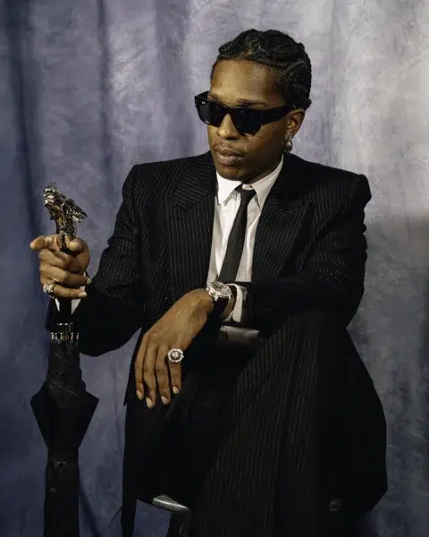 A$AP Rocky con un traje negro de rayas, gafas de sol Ray-Ban, sentado con un paraguas cuyo mango esculpido representa una cabeza de águila.