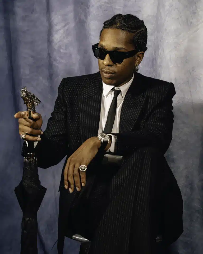 A$AP Rocky in a black striped suit, wearing Ray-Ban sunglasses, seated with an umbrella featuring a sculpted eagle-head handle.