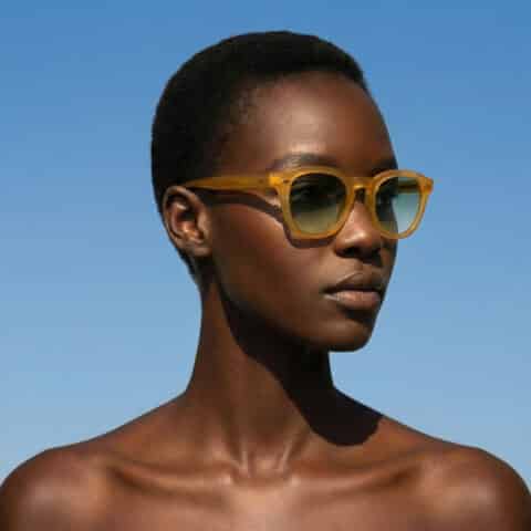 Portrait en extérieur d’un modèle portant les lunettes de soleil Dorata en acétate jaune miel transparent avec verres dégradés verts, ciel bleu en arrière-plan
