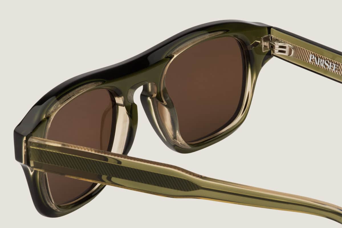 Primer plano de las gafas de sol Freschezza en acetato verde oliva translúcido con lentes marrones, detalle de bisagra y varillas estriadas sobre fondo beige claro