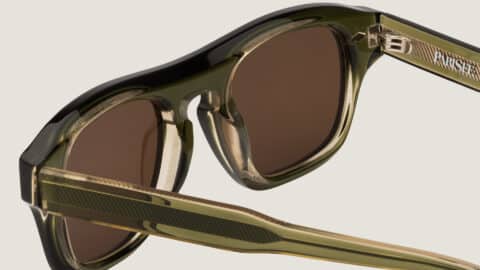 Primer plano de las gafas de sol Freschezza en acetato verde oliva translúcido con lentes marrones, detalle de bisagra y varillas estriadas sobre fondo beige claro