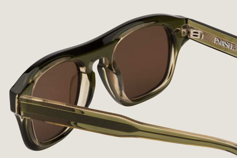 Gros plan des lunettes de soleil Freschezza en acétate vert olive translucide avec verres bruns, détail charnière et branches nervurées sur fond beige