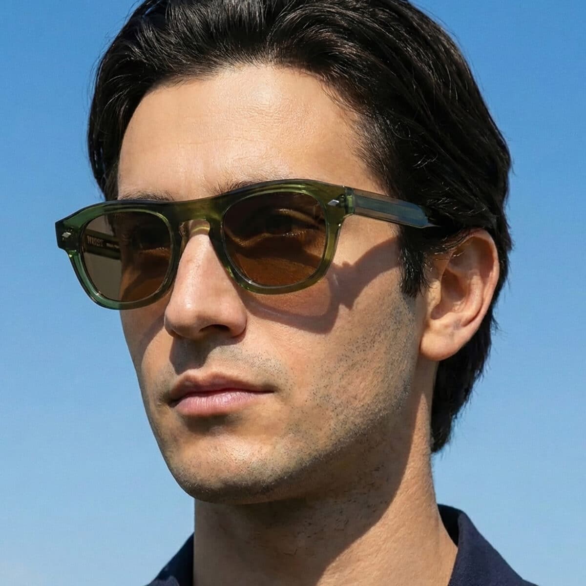 Retrato al aire libre de un hombre con las gafas de sol Freschezza en acetato verde oliva translúcido con lentes marrones, con cielo azul de fondo