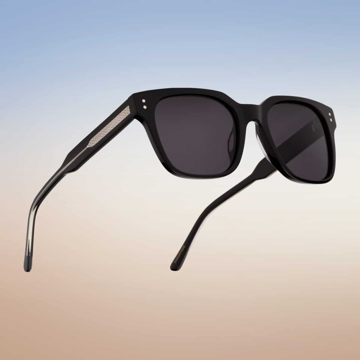 Vista lateral abierta de las gafas de sol Essentiel N1 en acetato negro mate con lentes gris oscuro, sobre fondo degradado cielo