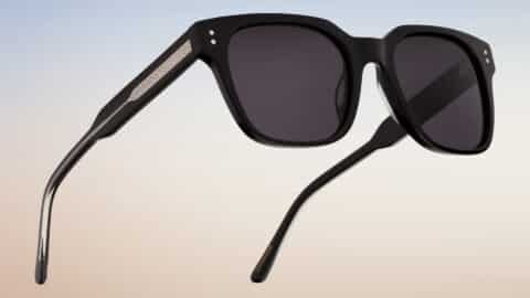 Vista lateral abierta de las gafas de sol Essentiel N1 en acetato negro mate con lentes gris oscuro, sobre fondo degradado cielo