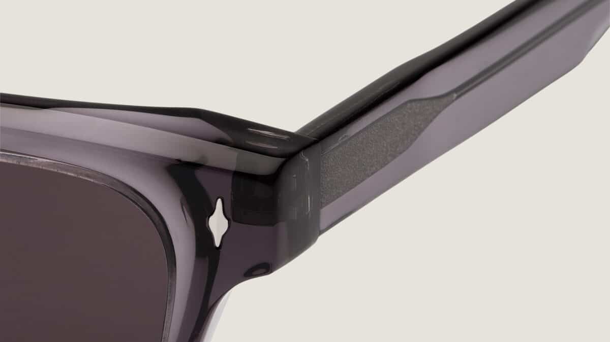 Detalle de la bisagra de las gafas de sol Essentiel N2 sobre fondo claro: acetato ahumado translúcido, inserto texturizado en la varilla, remache decorativo y lente gris oscuro