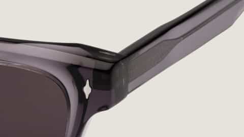 Detalle de la bisagra de las gafas de sol Essentiel N2 sobre fondo claro: acetato ahumado translúcido, inserto texturizado en la varilla, remache decorativo y lente gris oscuro