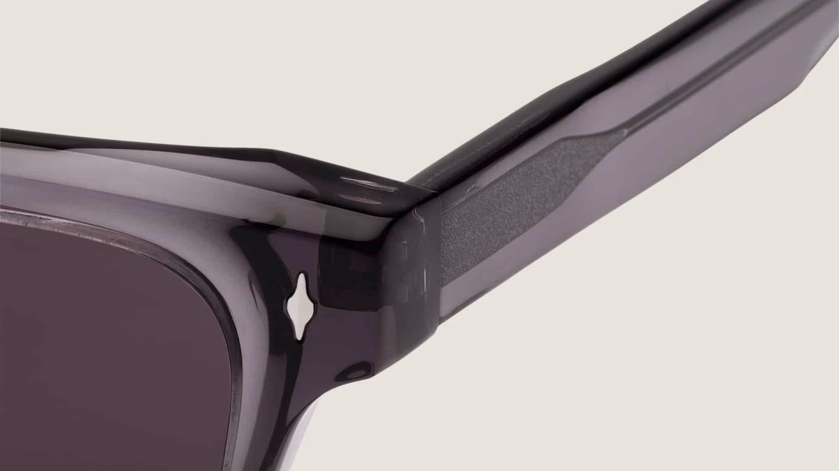 Detalle de la bisagra de las gafas de sol Essentiel N2 sobre fondo claro: acetato ahumado translúcido, inserto texturizado en la varilla, remache decorativo y lente gris oscuro